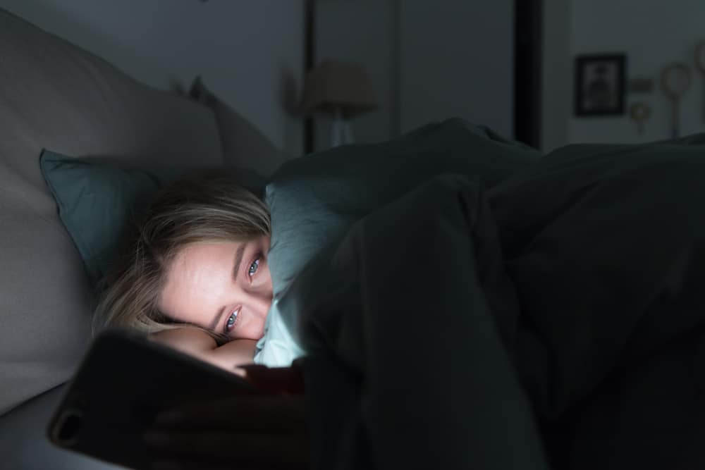 Portrait of young sleepy tired woman lying in bed under the blanket using smartphone at late night